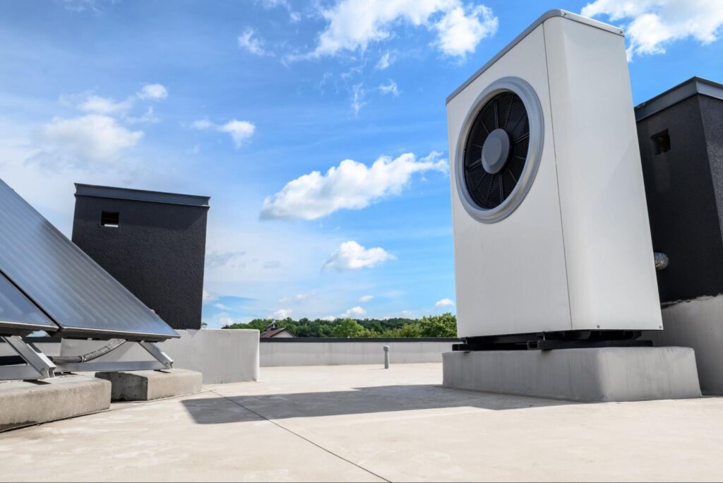 residential-cooling-wilmington-ma The rooftop scene showcases a sizable white HVAC unit beside solar panels and vents, all under a bright blue sky with scattered clouds. It's an ideal setup for residential cooling, efficiently managing the climate beneath with precision and care in Wilmington, MA.